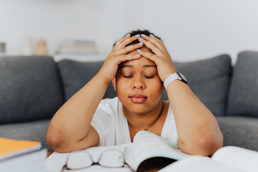 A woman with eyes closed holds her head in stress, surrounded by books in an indoor setting.